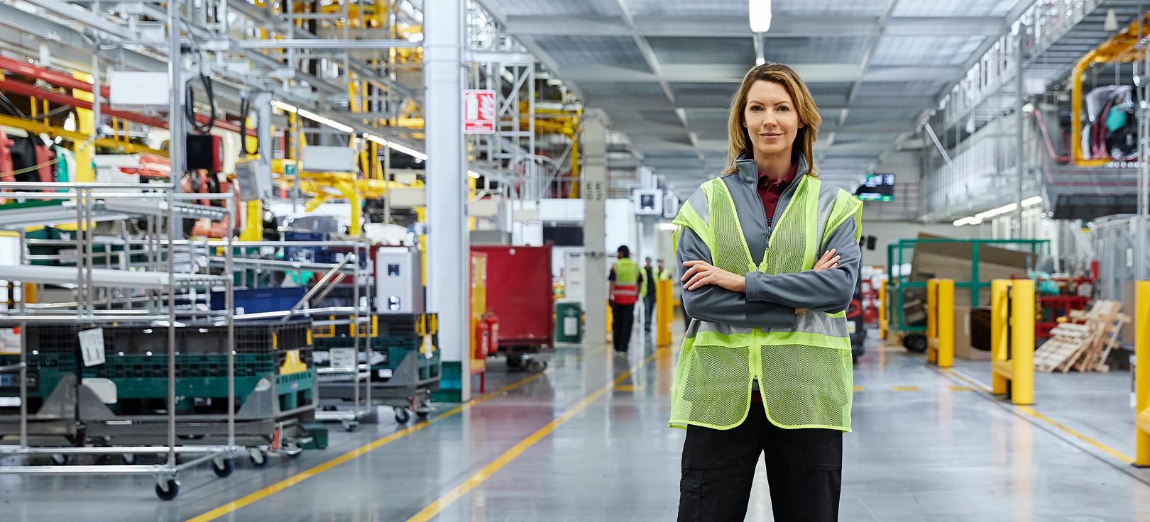 Lavoratore che indossa un giubbotto ad alta visibilità in piedi con le braccia incrociate in un capannone industriale circondato da attrezzature e macchinari industriali